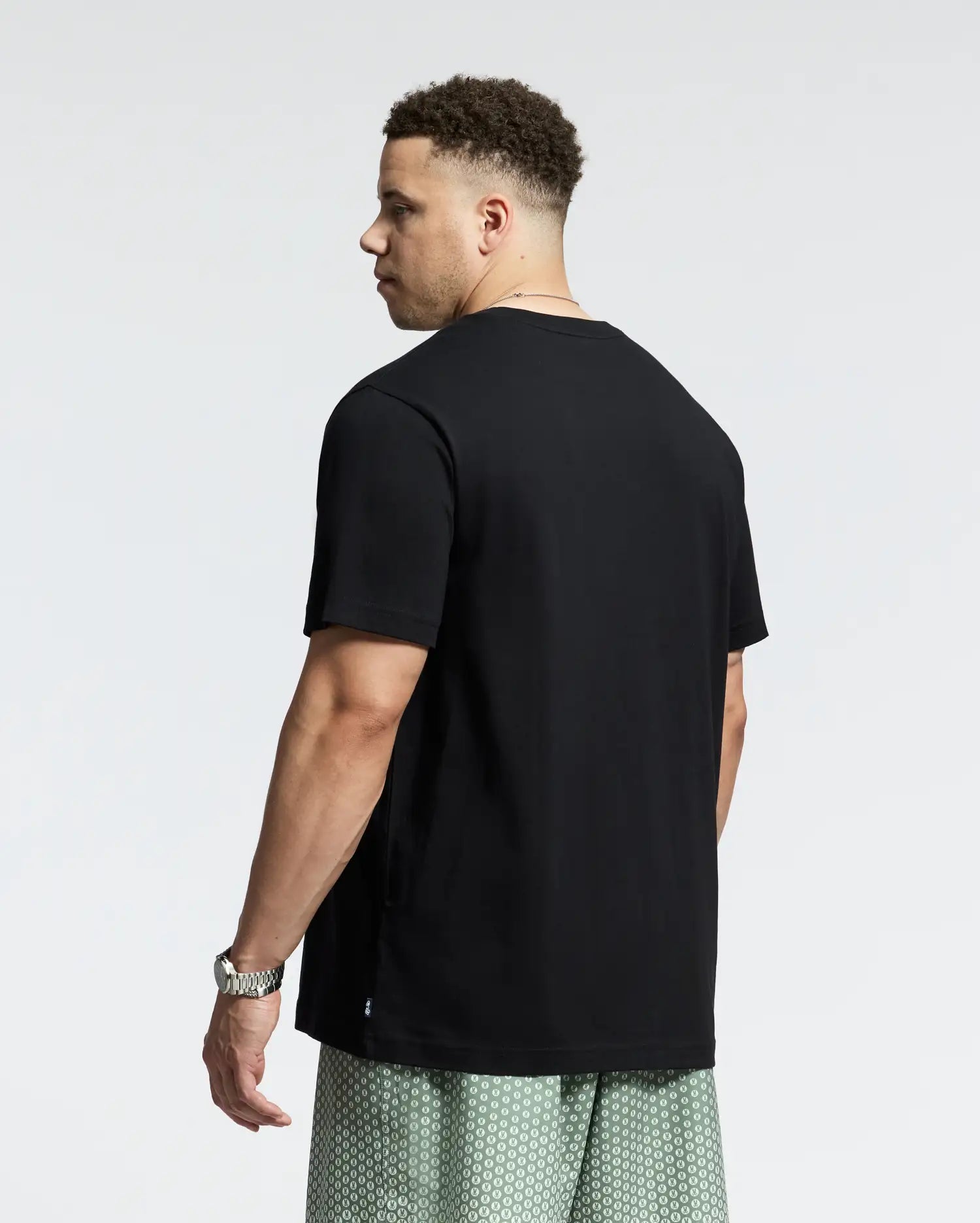 A man with short curly hair stands, glancing over his shoulder in a black Psycho Bunny Goldie Graphic Tee and patterned green shorts. A watch adorns his wrist, and the minimalistic white backdrop accentuates his cool style.