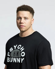 A curly-haired person with a neutral expression wears the Psycho Bunny Men's Big and Tall Goldie Graphic Tee (B9U606F200) in premium Pima cotton. This black t-shirt features the iconic Bunny graphic logo against a plain white backdrop.