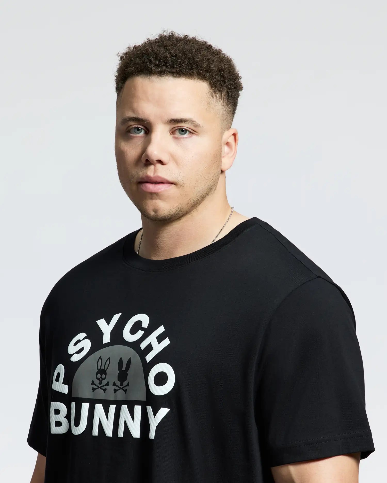 A curly-haired person with a neutral expression wears the Psycho Bunny Men's Big and Tall Goldie Graphic Tee (B9U606F200) in premium Pima cotton. This black t-shirt features the iconic Bunny graphic logo against a plain white backdrop.
