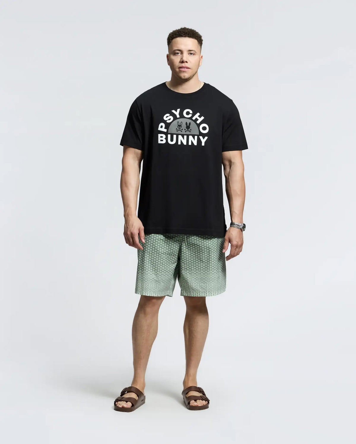 A man stands casually in a Psycho Bunny MENS BIG AND TALL GOLDIE GRAPHIC TEE (B9U606F200) made of black Pima cotton, featuring the iconic bunny graphic. He pairs it with light green patterned shorts, brown sandals, and a watch against a white background.