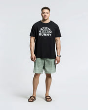 A man stands casually in a Psycho Bunny MENS BIG AND TALL GOLDIE GRAPHIC TEE (B9U606F200) made of black Pima cotton, featuring the iconic bunny graphic. He pairs it with light green patterned shorts, brown sandals, and a watch against a white background.