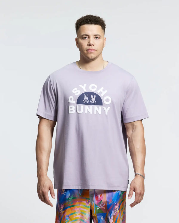 Wearing the Psycho Bunny MENS BIG AND TALL GOLDIE GRAPHIC TEE in lavender, made from soft Pima cotton, with two bunny graphics and vibrant patterned shorts, a person exudes casual elegance against a plain light background.