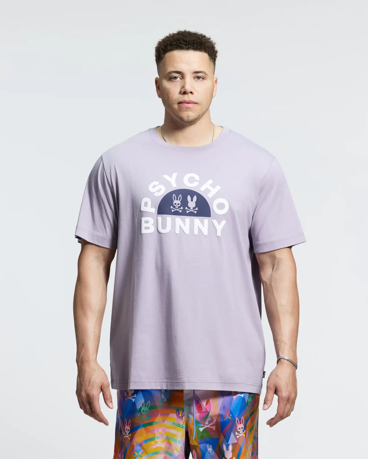 Wearing the Psycho Bunny MENS BIG AND TALL GOLDIE GRAPHIC TEE in lavender, made from soft Pima cotton, with two bunny graphics and vibrant patterned shorts, a person exudes casual elegance against a plain light background.