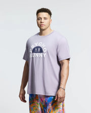A person wears a lavender Pima cotton tee, the Psycho Bunny Goldie Graphic Tee, with "Psycho Bunny" text and a bunny graphic. They pair it with vibrant shorts featuring abstract patterns on a light background for a lively ensemble.