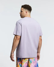 A person with short curly hair stands facing away, wearing a light purple Psycho Bunny Mens Big and Tall Goldie Graphic Tee - B9U606F200 adorned with a bunny graphic and colorful floral-patterned shorts. The plain bright background highlights this casual ensemble crafted from soft pima cotton.