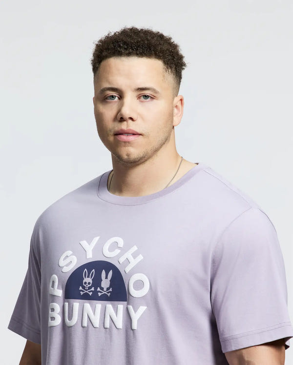A man is wearing the Psycho Bunny Men's Big and Tall Goldie Graphic Tee (B9U606F200) in light purple Pima cotton, featuring "Psycho Bunny" and bunny logos, against a plain background.