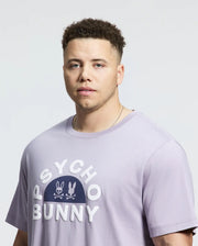 A man is wearing the Psycho Bunny Men's Big and Tall Goldie Graphic Tee (B9U606F200) in light purple Pima cotton, featuring "Psycho Bunny" and bunny logos, against a plain background.