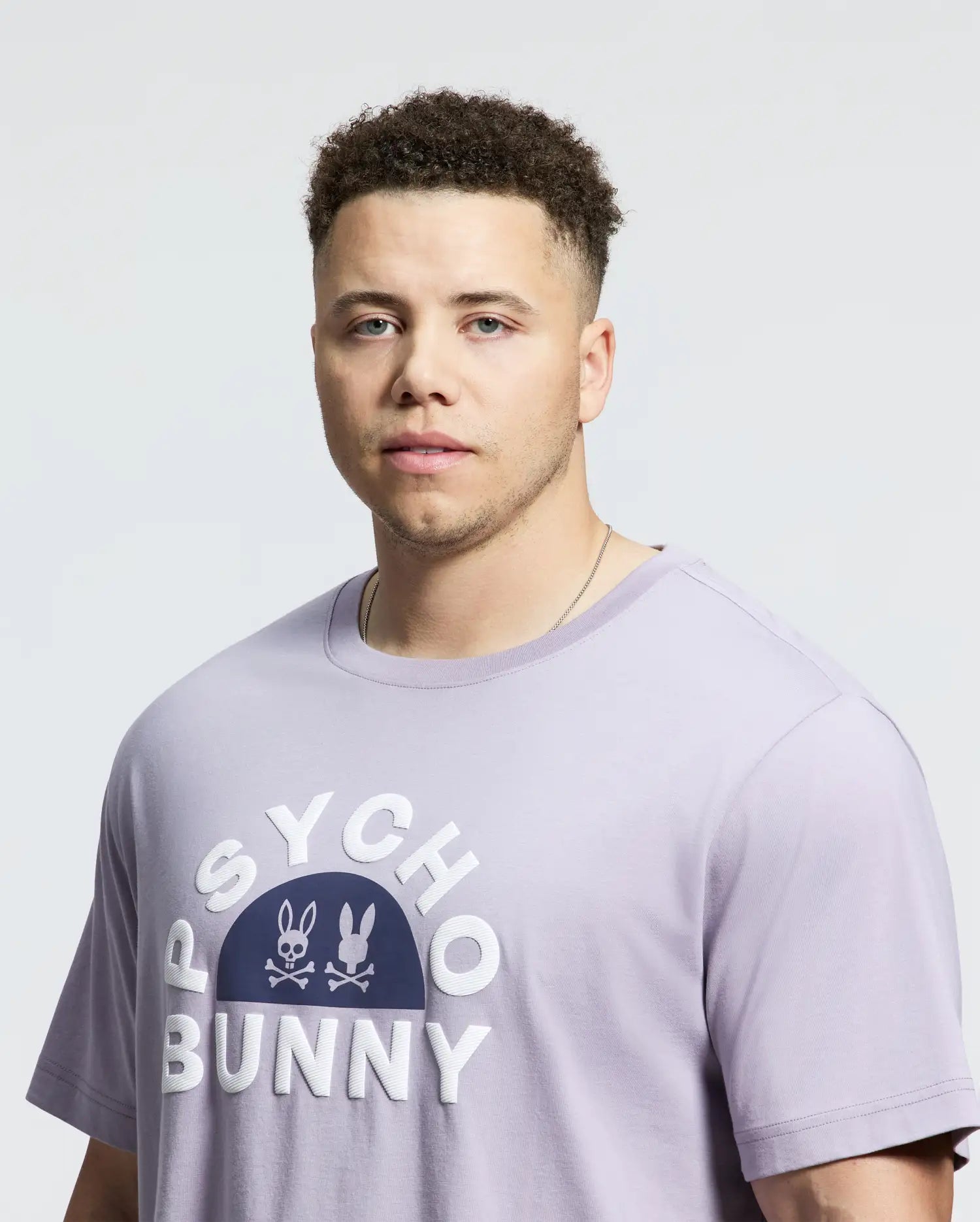 A man is wearing the Psycho Bunny Men's Big and Tall Goldie Graphic Tee (B9U606F200) in light purple Pima cotton, featuring "Psycho Bunny" and bunny logos, against a plain background.
