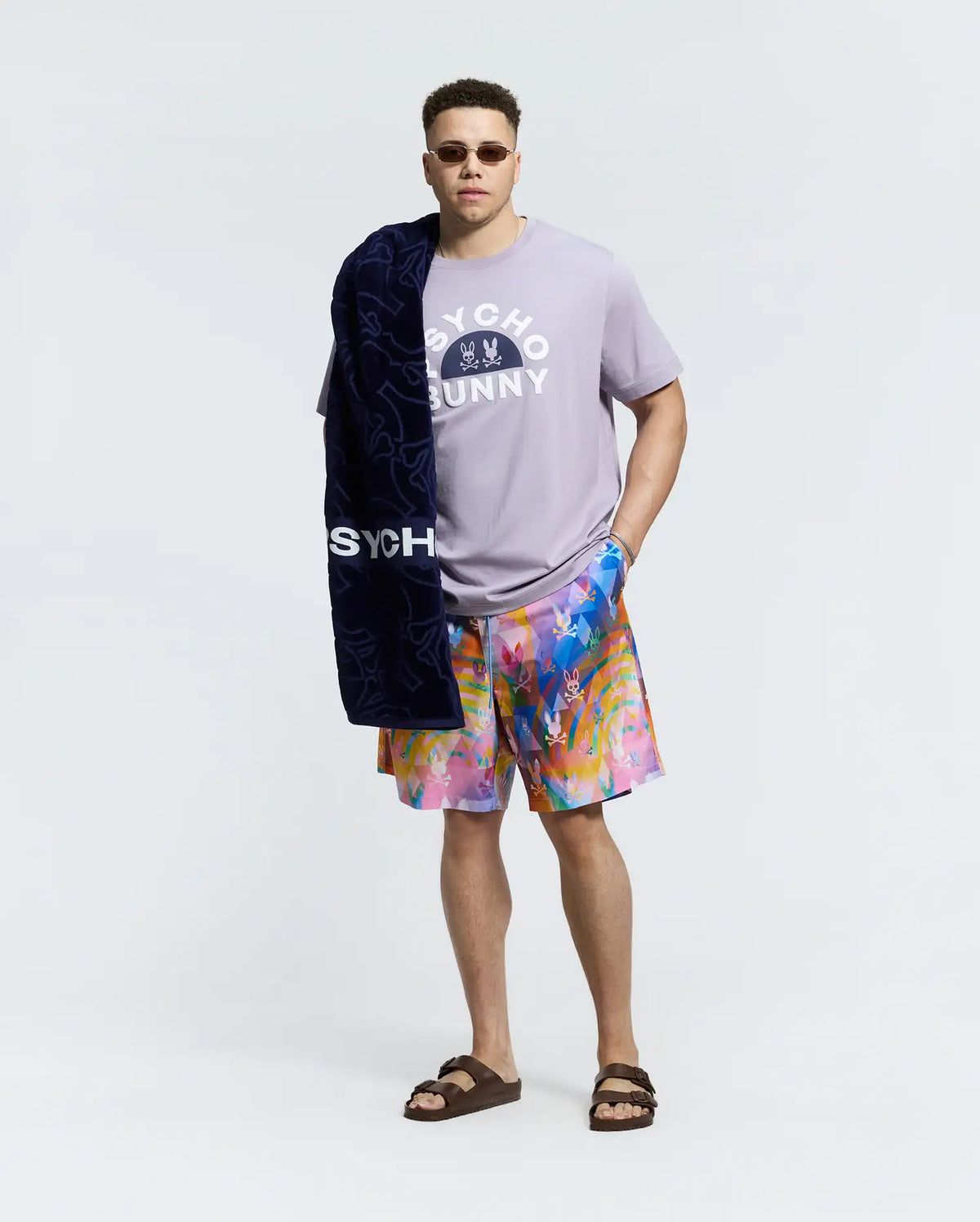 A man wears a lavender Psycho Bunny MENS BIG AND TALL GOLDIE GRAPHIC TEE with a bunny graphic, teamed with colorful shorts and brown sandals. Draped over his shoulder is a navy towel labeled "Psycho Bunny," while his sunglasses enhance the cool vibe against the plain white background.