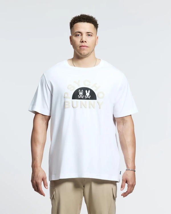 A person stands forward wearing a men's Big and Tall Goldie Graphic Tee in soft Peruvian Pima cotton with "PSYCHO BUNNY" logo and bunny graphic, paired with beige pants. The background is plain and light.