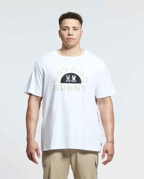 A person stands forward wearing a men's Big and Tall Goldie Graphic Tee in soft Peruvian Pima cotton with "PSYCHO BUNNY" logo and bunny graphic, paired with beige pants. The background is plain and light.