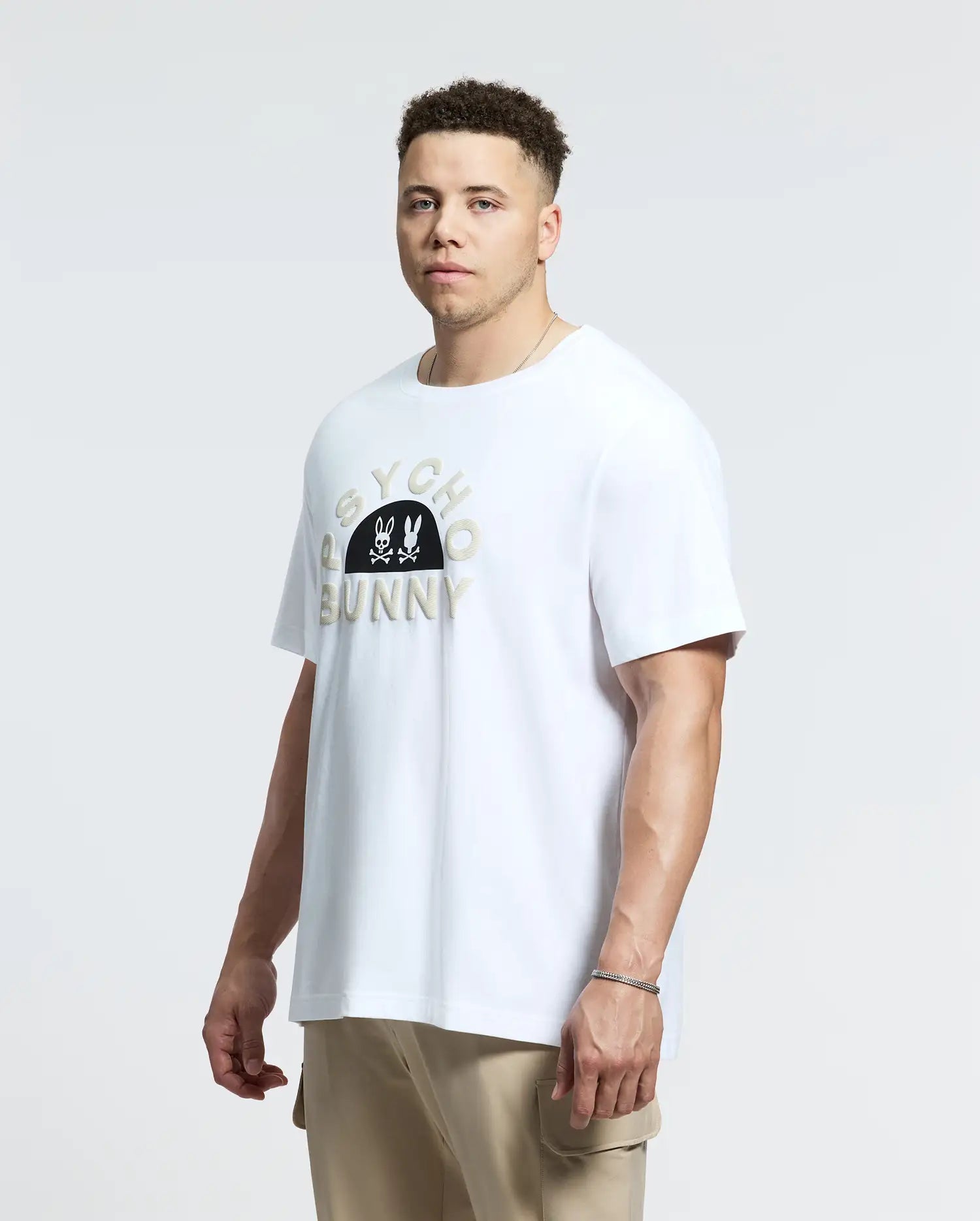 A man with short curly hair and beige pants wears a Psycho Bunny MENS BIG AND TALL GOLDIE GRAPHIC TEE - B9U606F200, crafted from luxurious Peruvian Pima cotton with the iconic bunny and crossbones graphic, set against a plain white background.