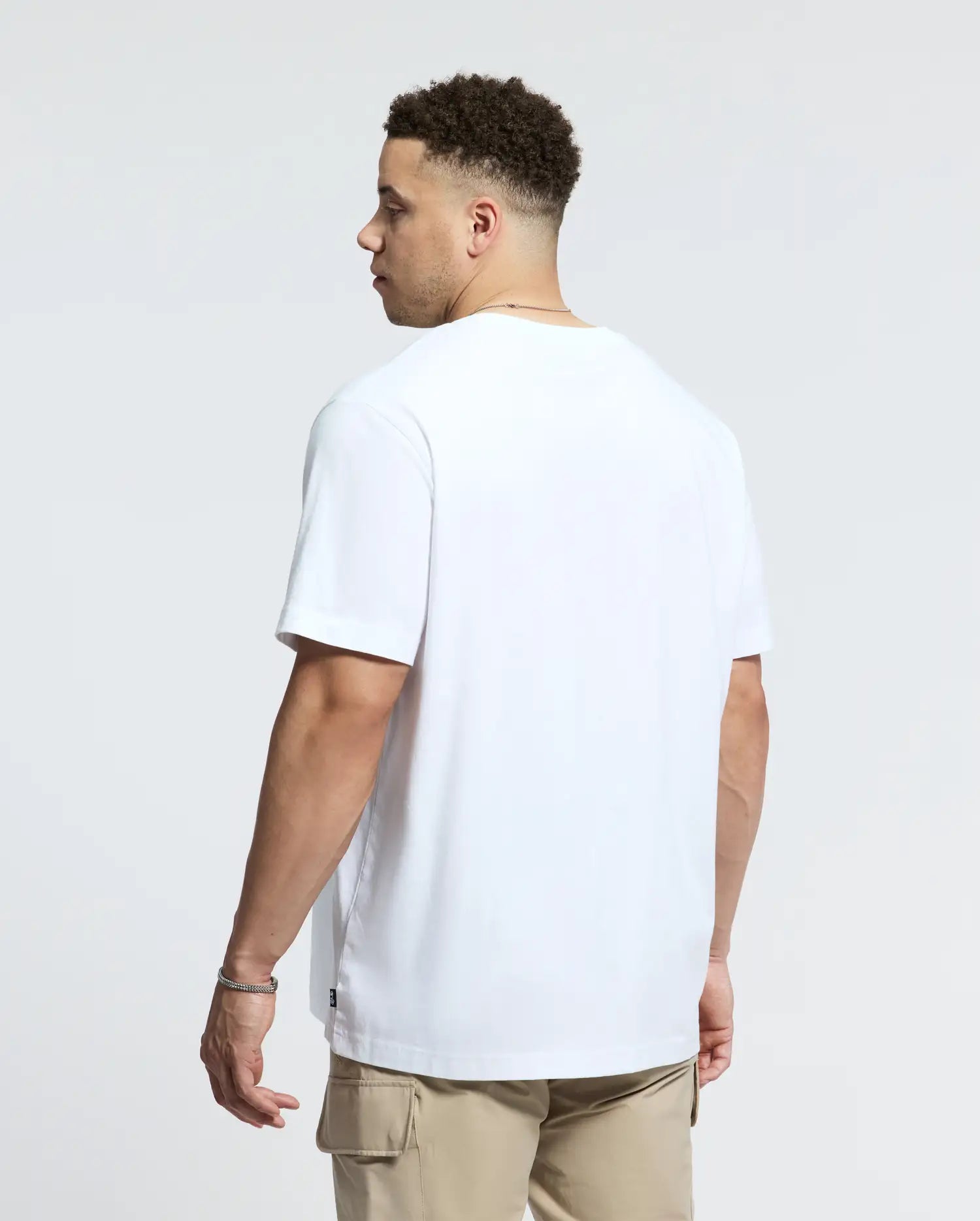 A person with short curly hair wears a Psycho Bunny Big and Tall Goldie Graphic Tee made from soft Peruvian Pima cotton, pairing it with beige cargo pants while gazing sideways against a plain light-colored background.