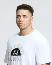A person with short curly hair wears a white Psycho Bunny Big and Tall Goldie Graphic Tee, made from Peruvian Pima cotton. The iconic graphic of two bunnies and a skull stands out as they gaze at the camera against a plain light background with a neutral expression.