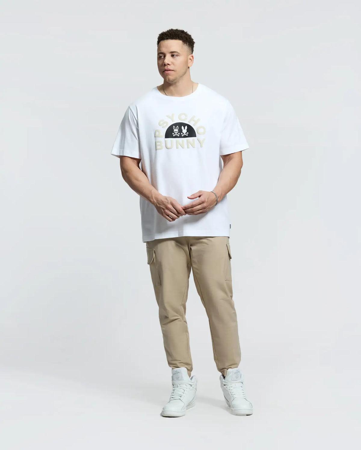 A person in a Psycho Bunny Goldie Graphic Tee (B9U606F200) with cartoon bunnies, beige joggers, and white sneakers stands against a plain white backdrop. The white T-shirt features "PSYCHO BUNNY" printed on it, made from premium Peruvian Pima cotton for ultimate comfort.
