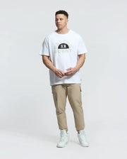A person in a Psycho Bunny Goldie Graphic Tee (B9U606F200) with cartoon bunnies, beige joggers, and white sneakers stands against a plain white backdrop. The white T-shirt features "PSYCHO BUNNY" printed on it, made from premium Peruvian Pima cotton for ultimate comfort.