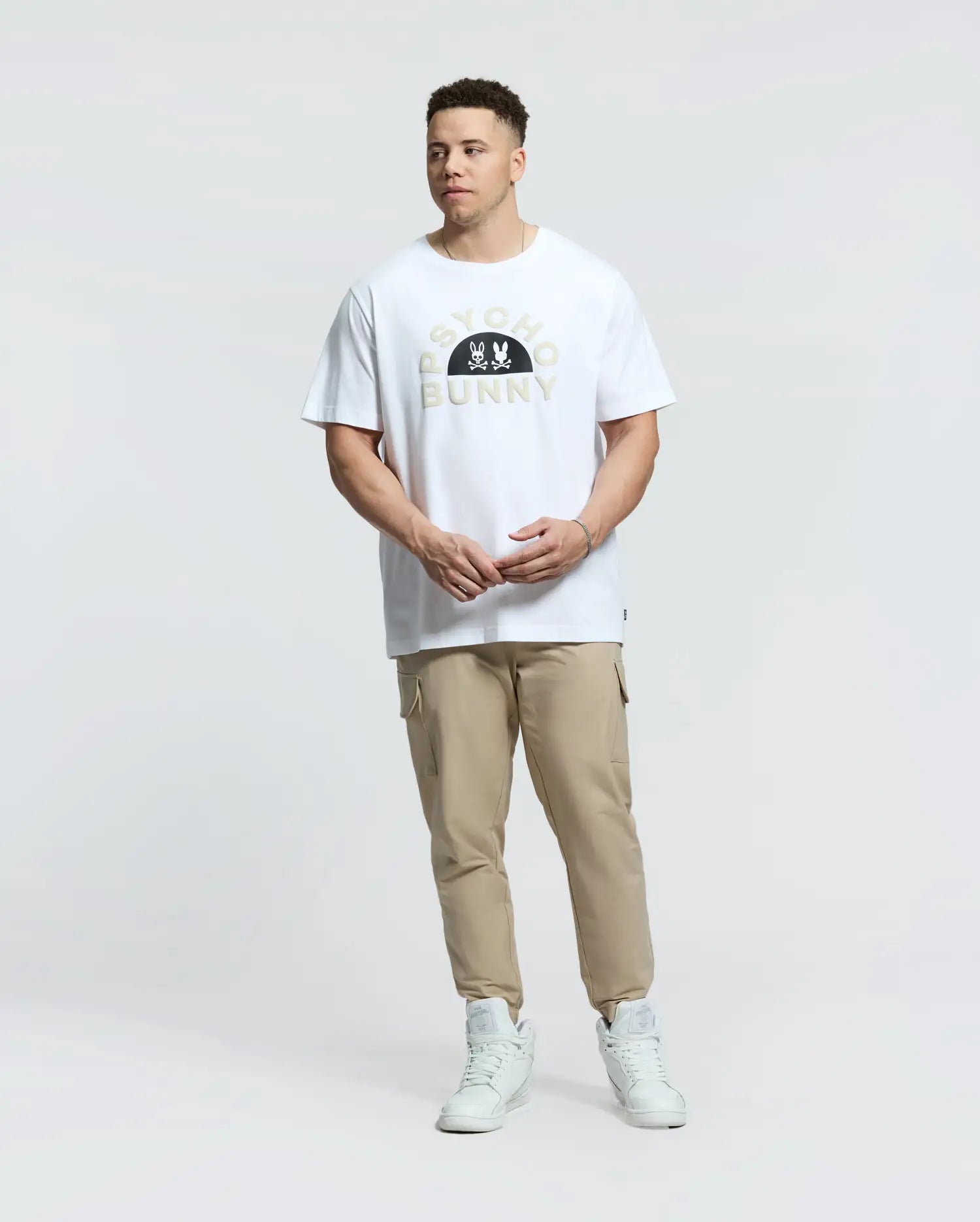 A person in a Psycho Bunny Goldie Graphic Tee (B9U606F200) with cartoon bunnies, beige joggers, and white sneakers stands against a plain white backdrop. The white T-shirt features "PSYCHO BUNNY" printed on it, made from premium Peruvian Pima cotton for ultimate comfort.