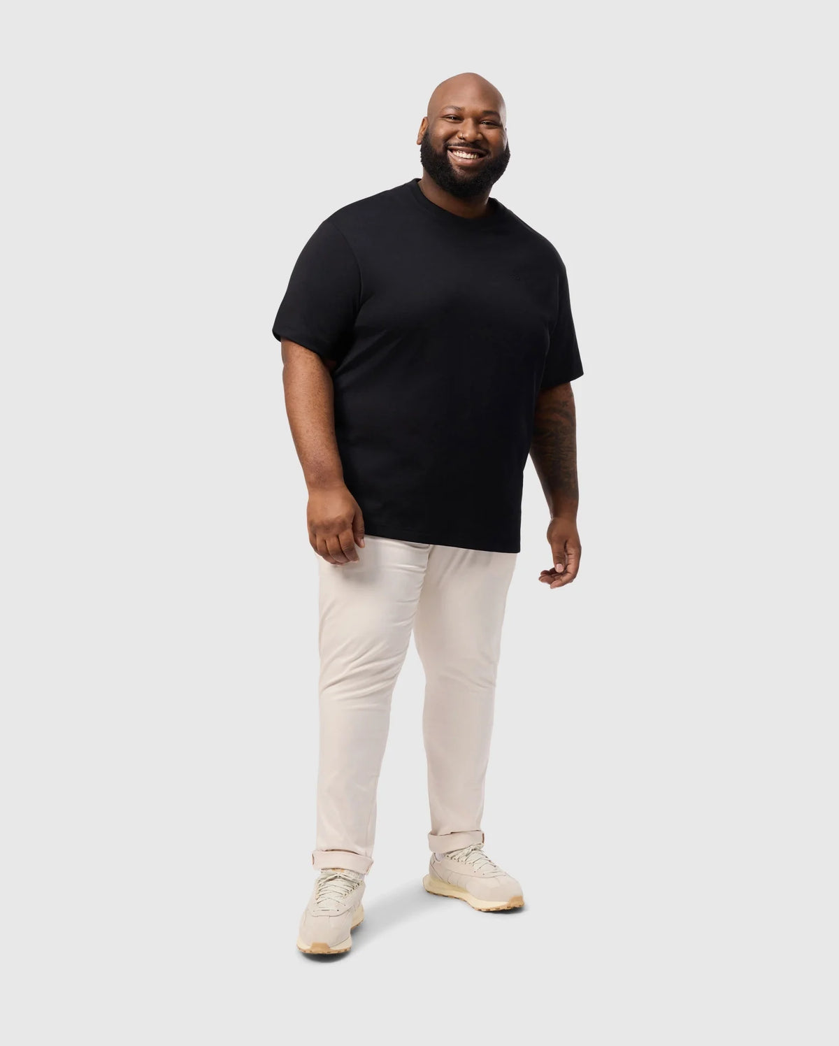 A smiling man stands against a plain background, wearing an oversized Psycho Bunny Duke tee, beige pants, and light-colored sneakers.