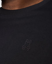 Close-up of a person wearing Psycho Bunny's BIG AND TALL DUKE OVERSIZED TEE in heavyweight Pima cotton, featuring a subtle embroidered chest bunny logo with crossbones on the black shirt.