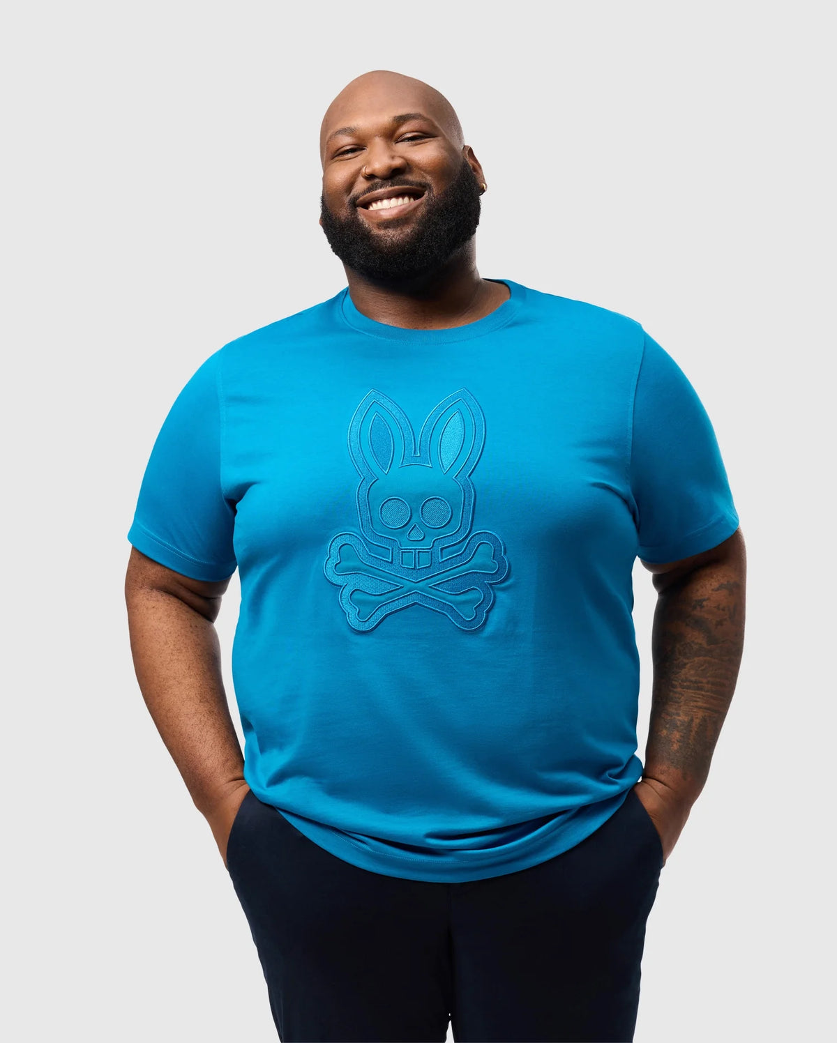 A man smiles, hands in pockets, wearing the Psycho Bunny BIG AND TALL MALTON GRAPHIC TEE - B9U795D200. The vibrant bunny and crossbones design pops against the light gray backdrop. Crafted with Peruvian artistry, it's made from soft Pima cotton.