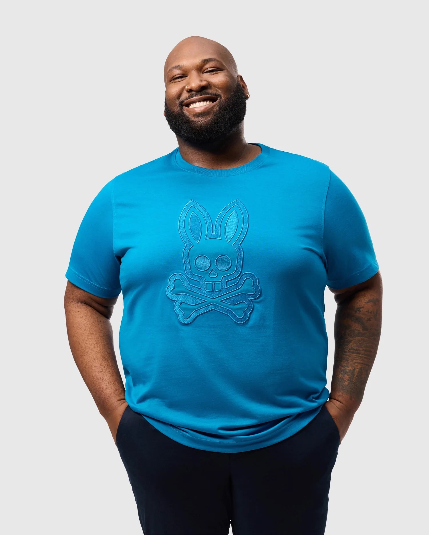 A man smiles, hands in pockets, wearing the Psycho Bunny BIG AND TALL MALTON GRAPHIC TEE - B9U795D200. The vibrant bunny and crossbones design pops against the light gray backdrop. Crafted with Peruvian artistry, it's made from soft Pima cotton.