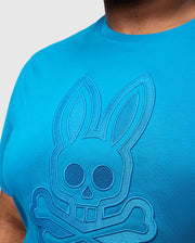 Wearing Psycho Bunny's BIG AND TALL MALTON GRAPHIC TEE, a person dons a bright blue Pima cotton shirt featuring an embossed rabbit skull with crossbones. Elongated ears and stitched outlines create a 3D effect that showcases exquisite Peruvian craftsmanship on the upper part of the tee.