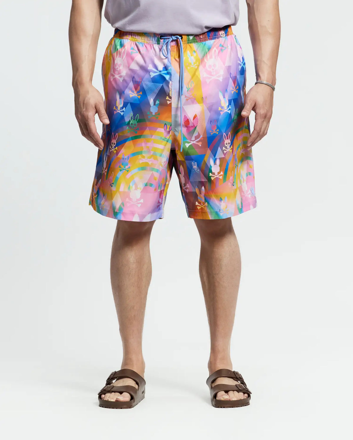 Wearing Psycho Bunny's MENS BIG AND TALL MILOS SWIM TRUNK - B9W446F200 with a bright abstract floral design, the person pairs it with a gray shirt and brown sandals, standing against a plain white backdrop.
