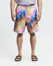Wearing Psycho Bunny's MENS BIG AND TALL MILOS SWIM TRUNK - B9W446F200 with a bright abstract floral design, the person pairs it with a gray shirt and brown sandals, standing against a plain white backdrop.