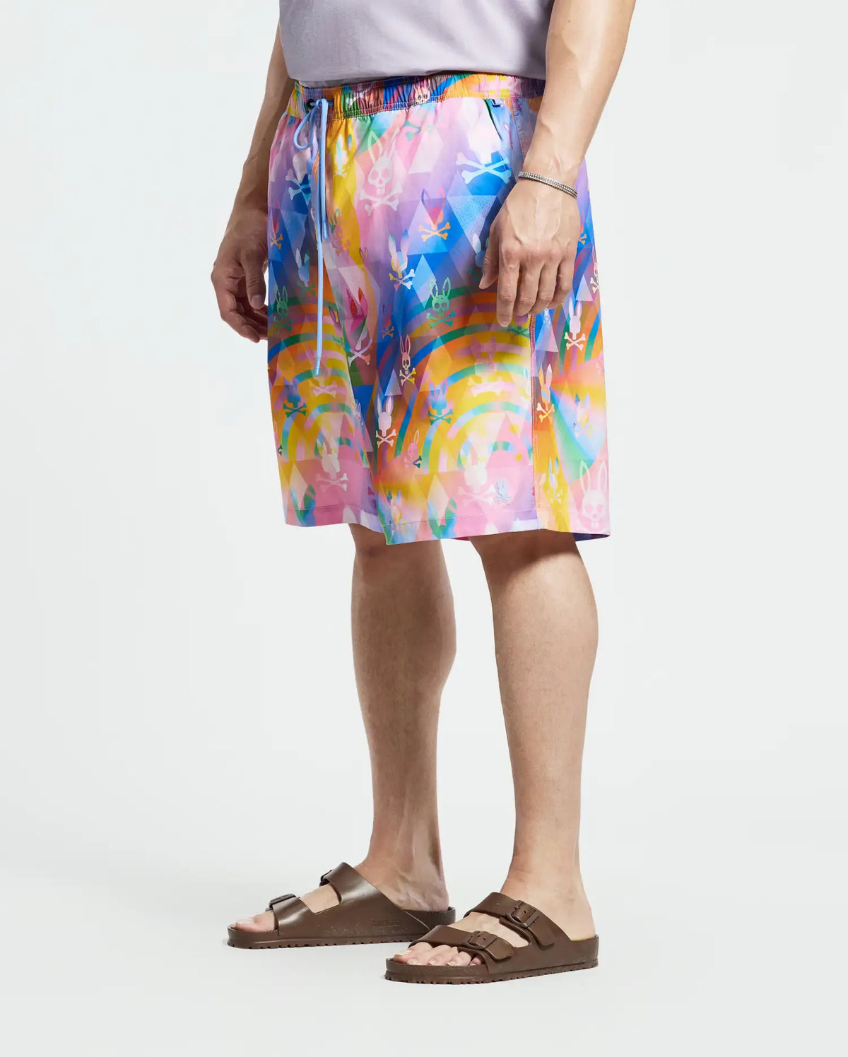 A person wears Psycho Bunny's MENS BIG AND TALL MILOS SWIM TRUNK - B9W446F200, featuring a vibrant abstract pattern in blues, pinks, and yellows with a drawstring waist and quick-dry fabric, paired with brown sandals.