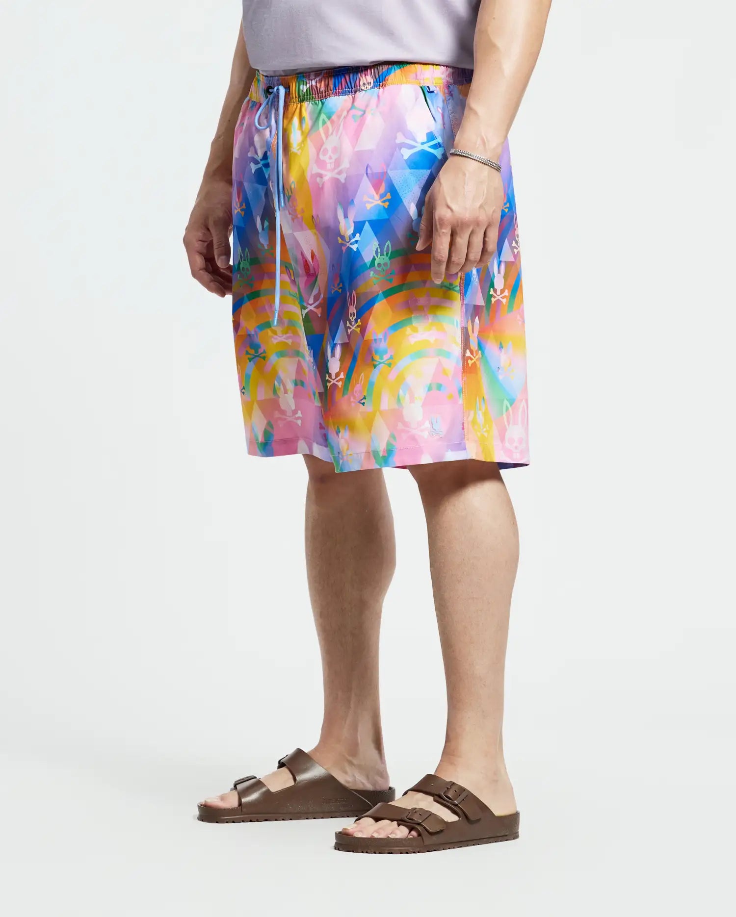 A person wears Psycho Bunny's MENS BIG AND TALL MILOS SWIM TRUNK - B9W446F200, featuring a vibrant abstract pattern in blues, pinks, and yellows with a drawstring waist and quick-dry fabric, paired with brown sandals.
