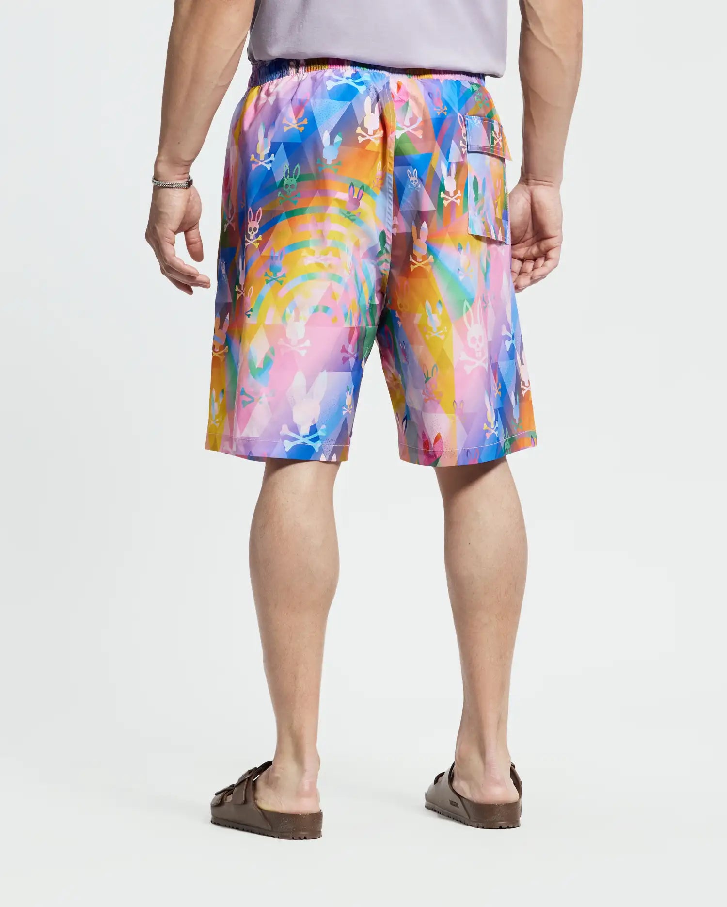 A person is shown from behind wearing a short-sleeved gray shirt, Psycho Bunny Big and Tall Milos Swim Trunk with a colorful abstract pattern of pink, blue, orange, and yellow in quick-dry fabric, and brown sandals.