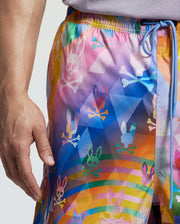 A person wearing Psycho Bunny's colorful Milos Swim Trunk (B9W446F200) with a pastel rainbow gradient and multi-colored bunny skull graphics. A light blue drawstring complements the quick-dry fabric, with their hand resting by their side.
