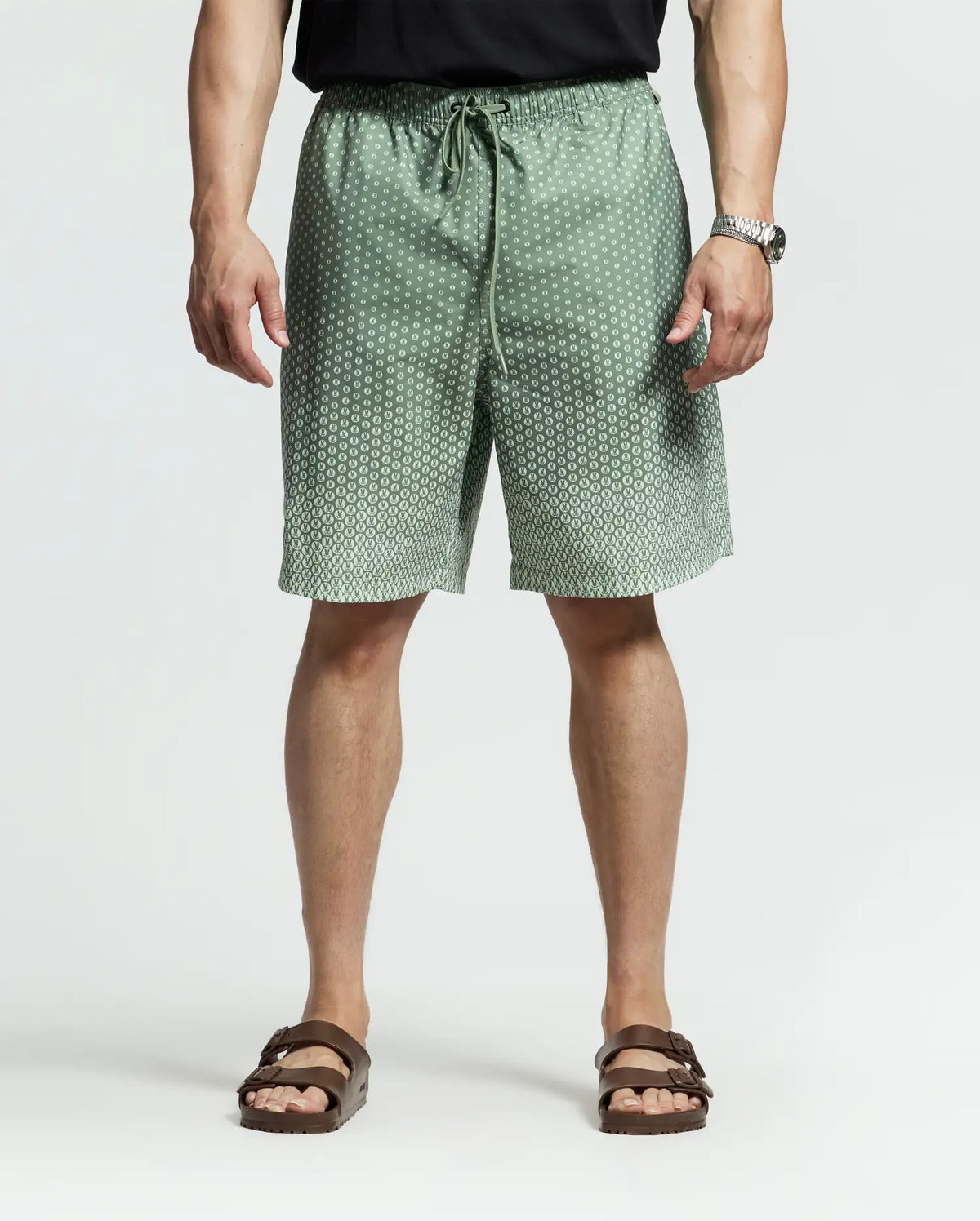 A person in Psycho Bunny's Big and Tall Bendigo Printed Swim Trunk featuring a subtle bunny pattern pairs it with a black shirt, brown sandals, and a watch on their left wrist, creating a laid-back travel style against a plain backdrop.