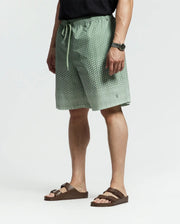 Leaning against a plain background, the person sports Psycho Bunny's green shorts with a tiny white polka dot pattern, styled in a relaxed jet-setting manner. They're wearing a black T-shirt, brown sandals, and have a black watch on their left wrist.