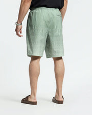 Facing away, they wear a black shirt and Psycho Bunny's Bendigo Printed Swim Trunk in green with a white dotted pattern, offering a relaxed jet-setting vibe. Brown sandals complete the look. The shorts have an elastic waistband for quick-dry comfort, and a watch adorns their left wrist against the plain white backdrop.