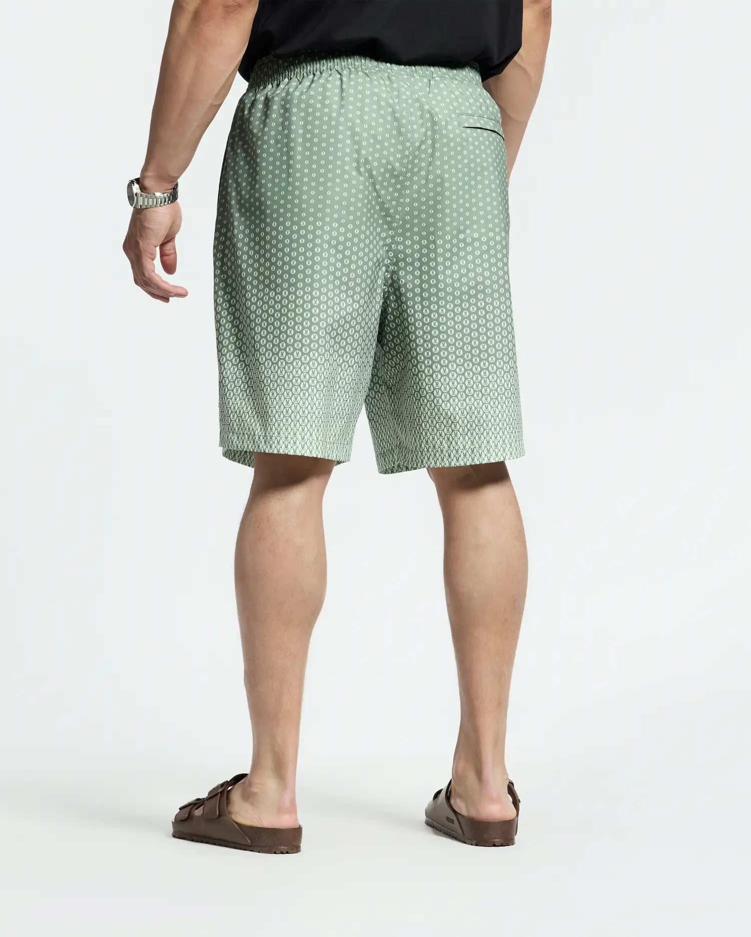 Facing away, they wear a black shirt and Psycho Bunny's Bendigo Printed Swim Trunk in green with a white dotted pattern, offering a relaxed jet-setting vibe. Brown sandals complete the look. The shorts have an elastic waistband for quick-dry comfort, and a watch adorns their left wrist against the plain white backdrop.