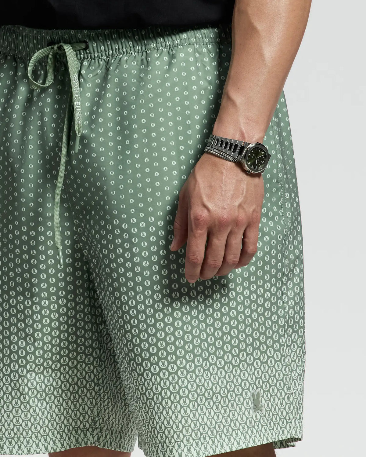 A person in light green Psycho Bunny MENS BIG AND TALL BENDIGO PRINTED SWIM TRUNK - B9W619F200 with a playful pattern stands with a relaxed jet-setting style, complemented by quick-dry fabric. They sport a silver wristwatch with a dark face against a plain gray background.