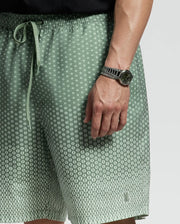 A person in light green Psycho Bunny MENS BIG AND TALL BENDIGO PRINTED SWIM TRUNK - B9W619F200 with a playful pattern stands with a relaxed jet-setting style, complemented by quick-dry fabric. They sport a silver wristwatch with a dark face against a plain gray background.