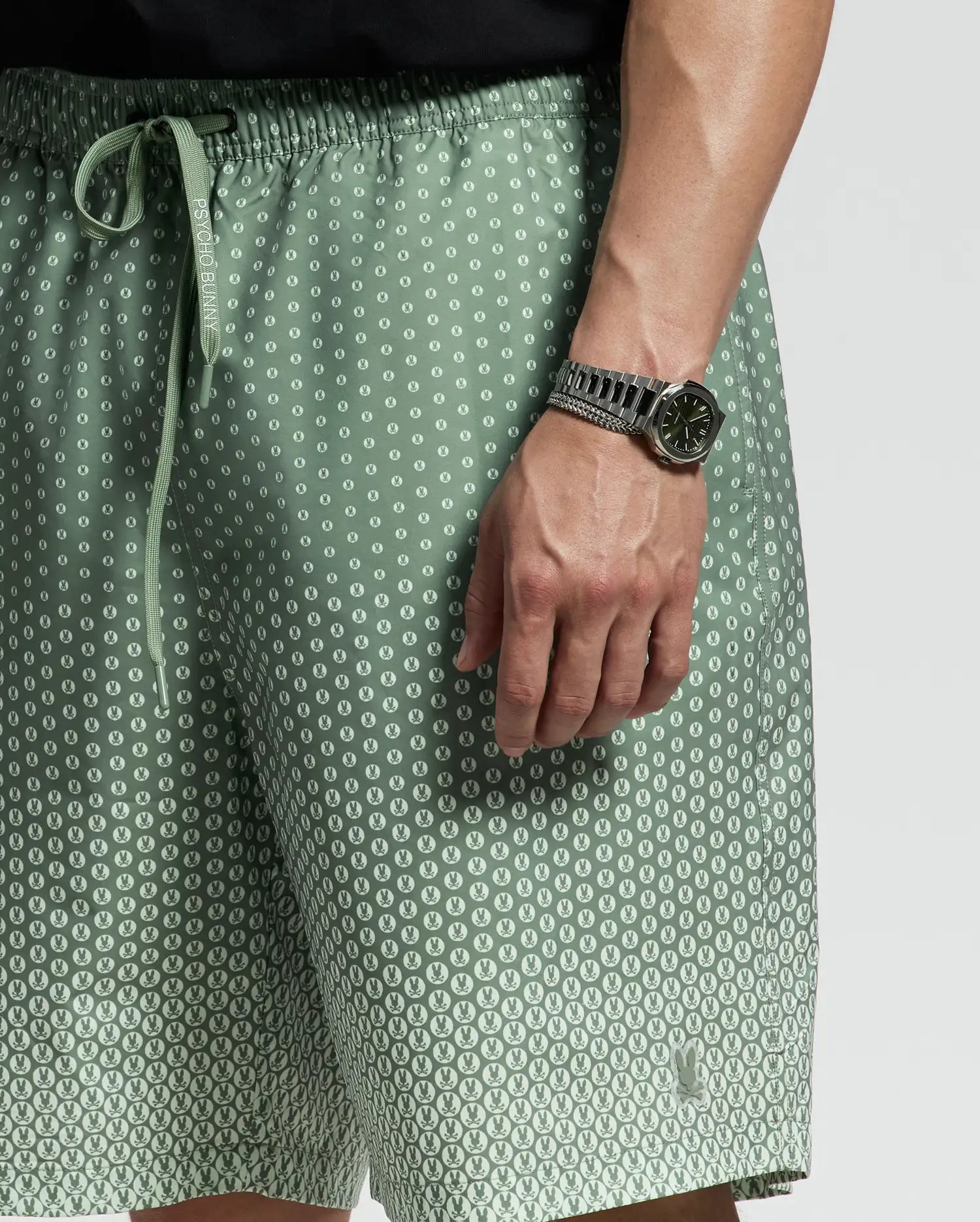 A person in light green Psycho Bunny MENS BIG AND TALL BENDIGO PRINTED SWIM TRUNK - B9W619F200 with a playful pattern stands with a relaxed jet-setting style, complemented by quick-dry fabric. They sport a silver wristwatch with a dark face against a plain gray background.