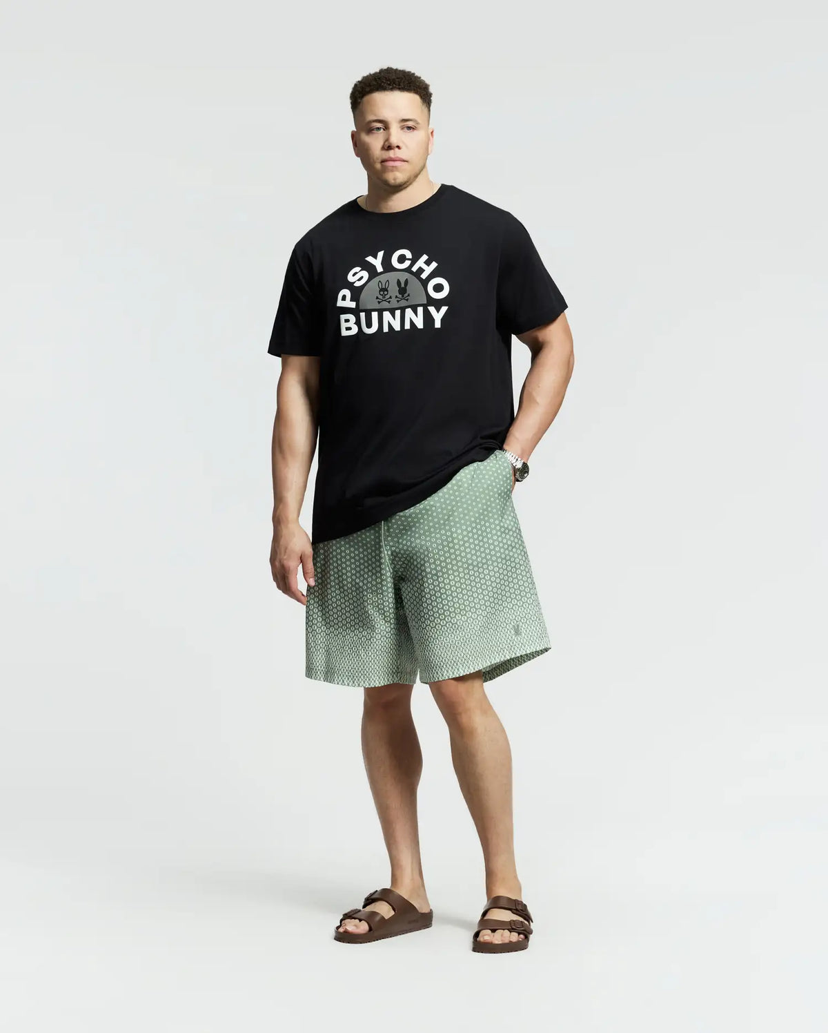A man exudes confidence in a black Psycho Bunny tee with the brand's signature pattern, paired with light green MENS BIG AND TALL BENDIGO PRINTED SWIM TRUNK shorts featuring quick-dry fabric for a laid-back travel style. His look is completed with brown sandals against a simple light gray backdrop.