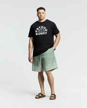 A man exudes confidence in a black Psycho Bunny tee with the brand's signature pattern, paired with light green MENS BIG AND TALL BENDIGO PRINTED SWIM TRUNK shorts featuring quick-dry fabric for a laid-back travel style. His look is completed with brown sandals against a simple light gray backdrop.