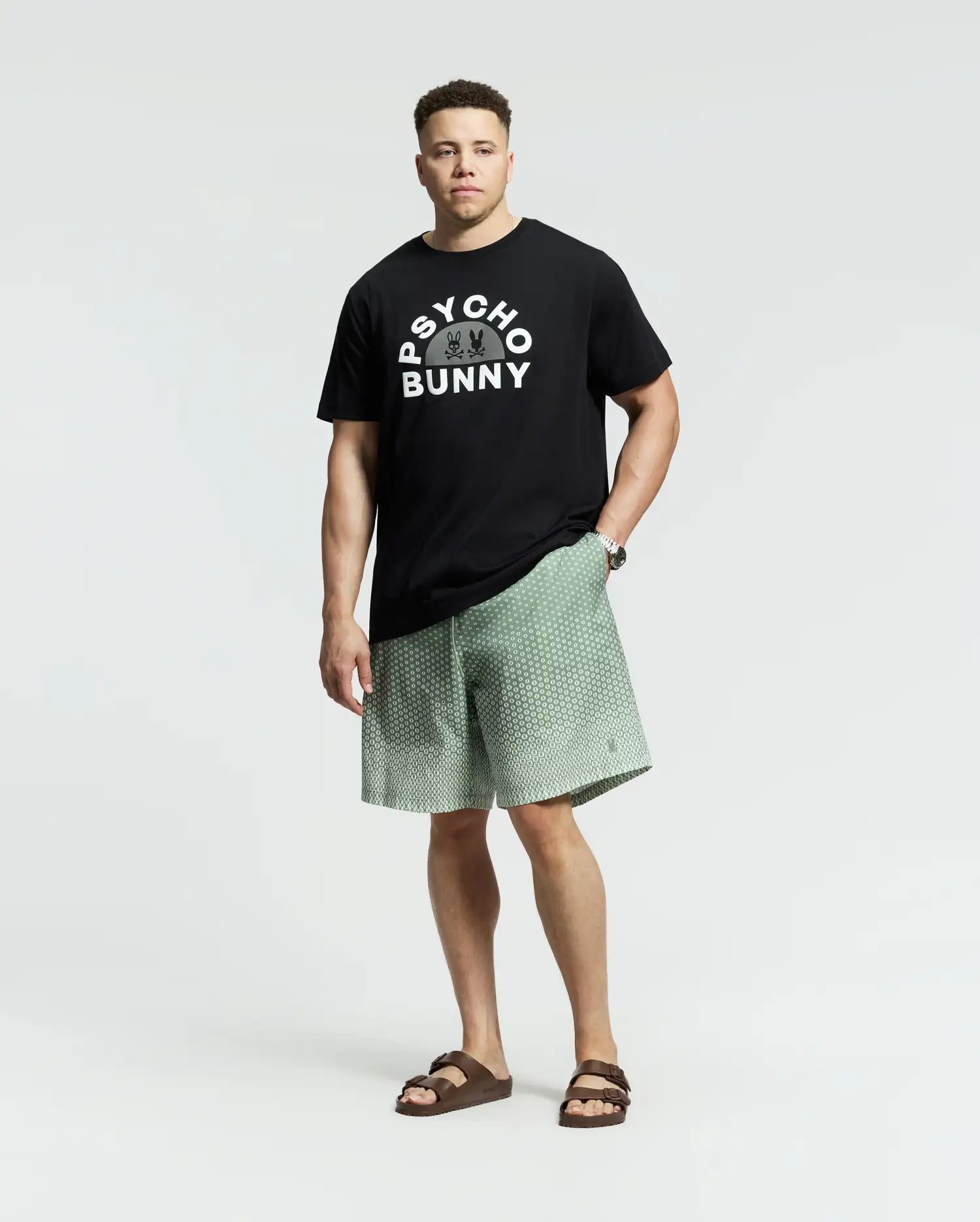 A man exudes confidence in a black Psycho Bunny tee with the brand's signature pattern, paired with light green MENS BIG AND TALL BENDIGO PRINTED SWIM TRUNK shorts featuring quick-dry fabric for a laid-back travel style. His look is completed with brown sandals against a simple light gray backdrop.