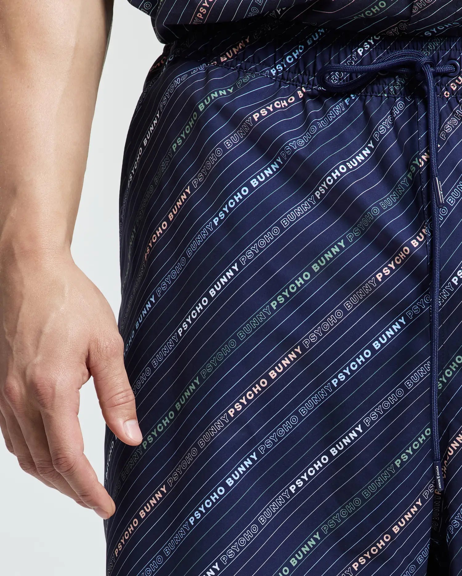 A person is wearing Psycho Bunny's blue MENS BIG AND TALL RENMARK PB PRINTED SWIM TRUNK with a quick-dry logo-patterned print. Their relaxed left hand rests by their side, while the drawstring peeks at the waist, ideal for laid-back beach days.