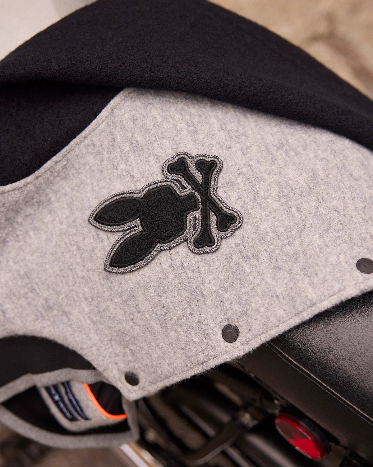 Close-up of a gray fabric on the Psycho Bunny MENS TOM WOOL BOMBER JACKET - B6J744D200, featuring a black emblem with a bunny and crossbones design. This elegant jacket's material is draped effortlessly over a motorcycle seat.