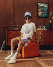 A young boy wearing the Kids Milos Bucket Reversible Hat from Psycho Bunny and trendy sunglasses sits on an orange suitcase. He's dressed in a white t-shirt, colorful shorts, and white sneakers. The room showcases a wooden cabinet, an embroidered vase with greenery, and a framed nautical painting.