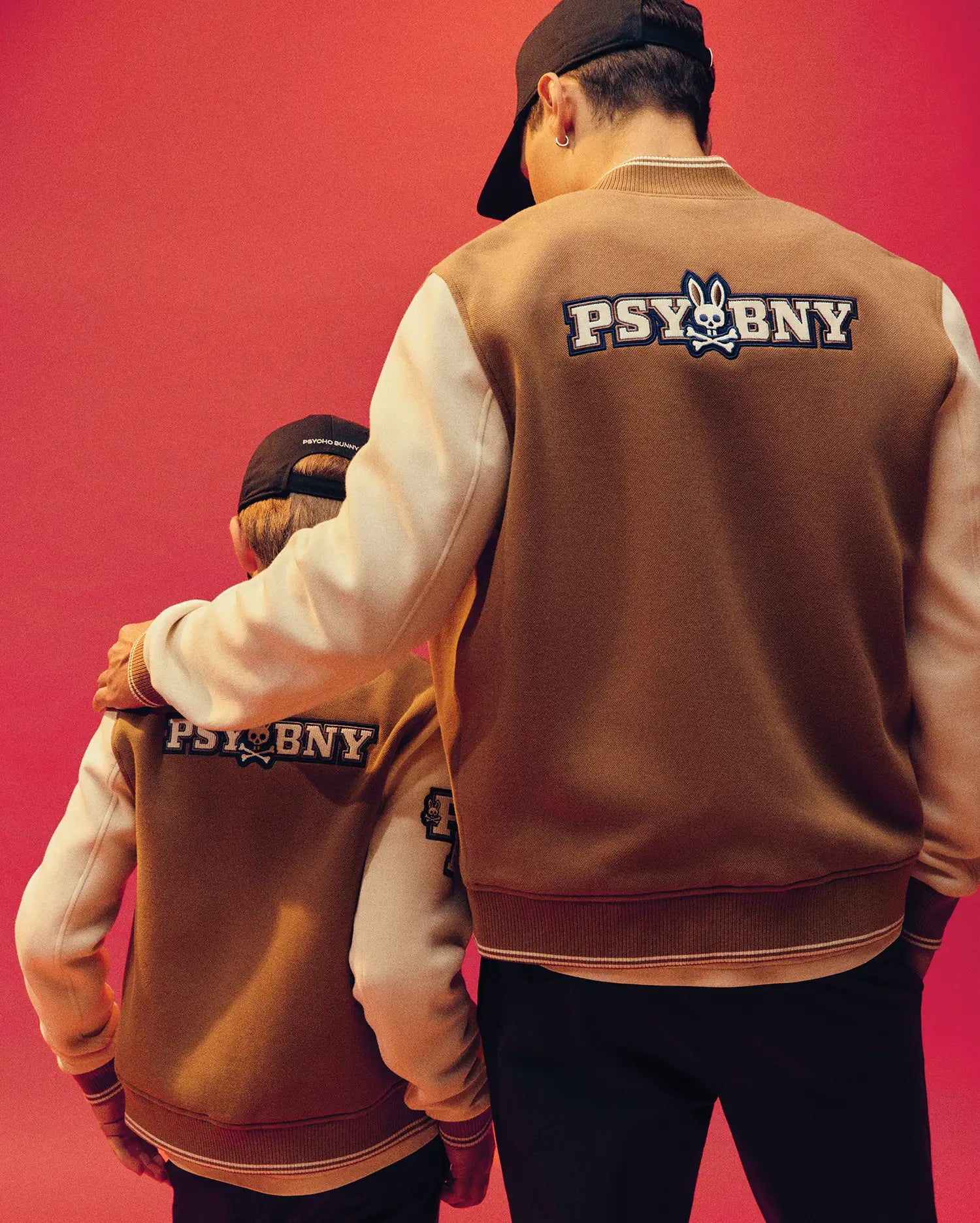 An adult and child stand side by side in matching Psycho Bunny MENS WADE KNIT BOMBER jackets (MO1100020), featuring white sleeves and an embroidered rabbit "PSYBUNY" logo on the back, set against a red background.