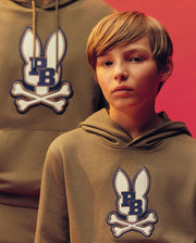 A young boy wears the Psycho Bunny KIDS WADE CORD PATCH HOODIE (KT0500031), a tan cotton blend hoodie with a blue and white bunny and crossbones patch, standing in front of another person in a similar hoodie against a red background.