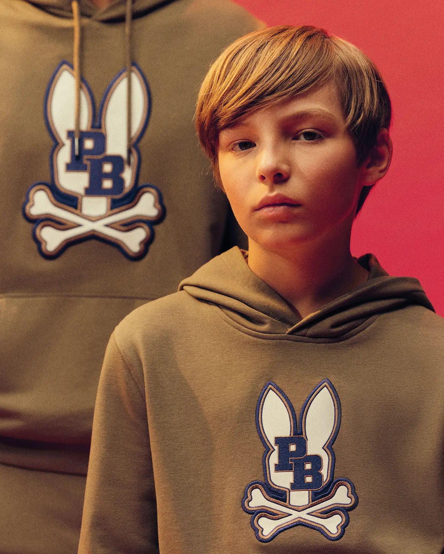 A young boy wears the Psycho Bunny KIDS WADE CORD PATCH HOODIE (KT0500031), a tan cotton blend hoodie with a blue and white bunny and crossbones patch, standing in front of another person in a similar hoodie against a red background.