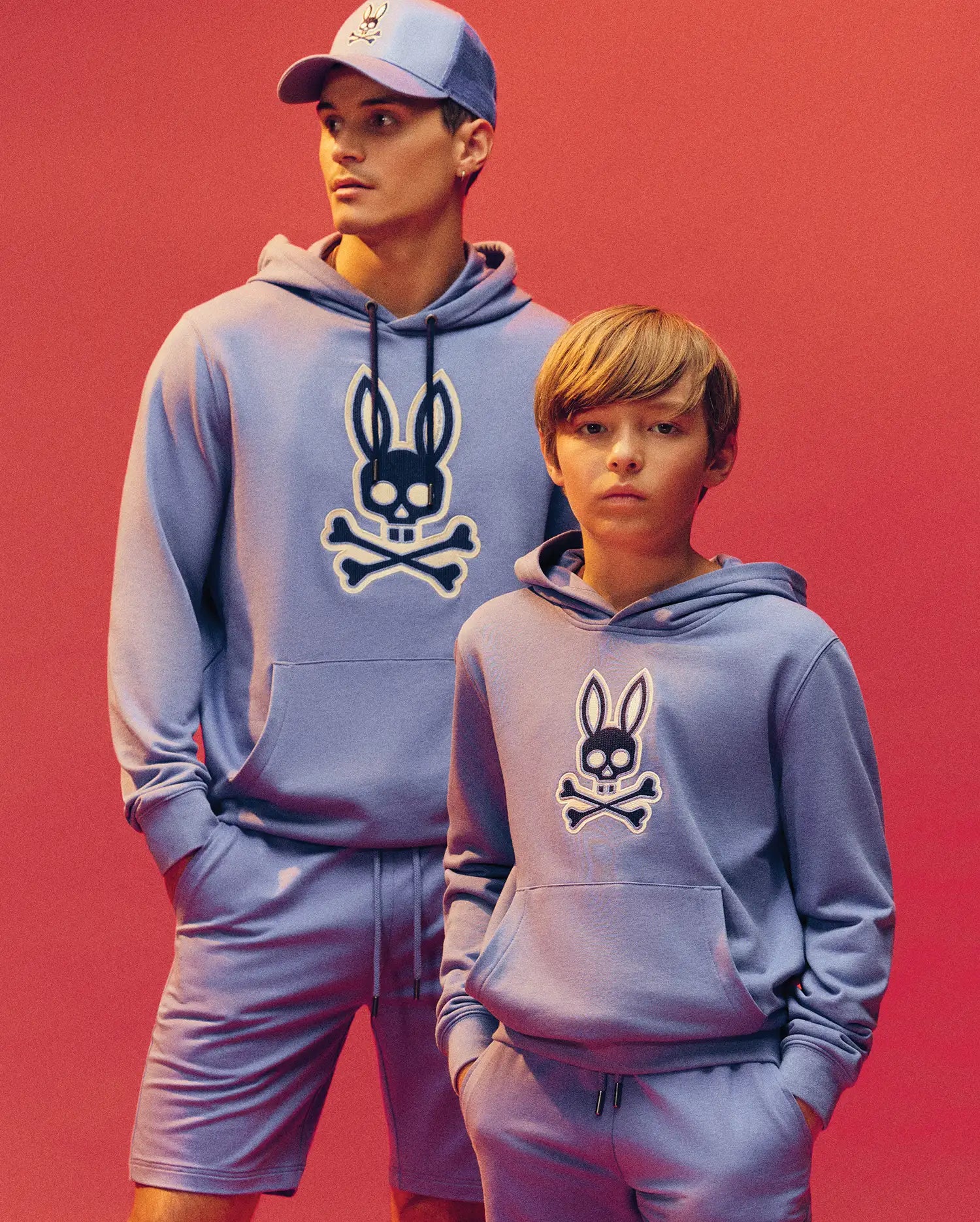 An adult and child wear matching lavender Psycho Bunny Ryder hoodies (KIDS RYDER BUNNY HOODIE - KT0500018) with an embroidered bunny patch featuring a black and white skull logo, posing together against a warm pink background.