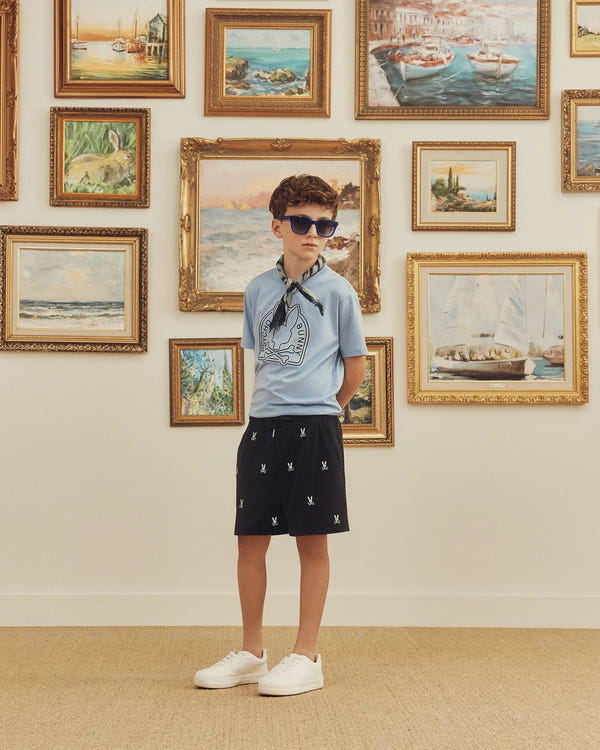 A child wearing sunglasses, the Psycho Bunny Kids Melbourne Graphic Tee - B0U410F200 in light blue made from eco-friendly Peruvian Pima cotton, black shorts, and white sneakers stands in front of a wall with framed landscape paintings.